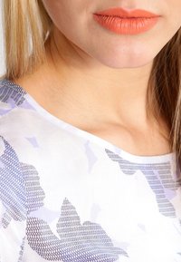 Chemisier blanc avec des motifs floraux et géométriques violets, col rond et texture lisse. Les lèvres du modèle sont colorées en orange clair.