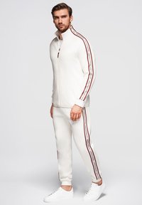 Chaqueta blanca con cremallera y pantalones a juego con rayas laterales marrones, confeccionados en un material suave y liso; combinados con zapatillas blancas.