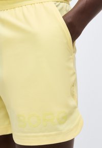Κοντινή λήψη κίτρινων σορτς με ελαστική μέση, πλαϊνή τσέπη και υφασματικό λογότυπο "BORG" κοντά στο τελείωμα στο χέρι ενός ατόμου με σκουρόχρωμο δέρμα.