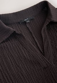 Gros plan sur un chemisier en tissu texturé foncé avec un col et une étiquette de marque "NEXT" visible à l'intérieur de l'encolure.
