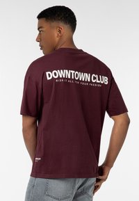 Bordeauxfarbenes T-Shirt mit kurzen Ärmeln, auf dem Rücken in Weiß "DOWNTOWN CLUB" und einem Slogan gedruckt, aus Baumwolle gefertigt und mit einem lockeren Schnitt.