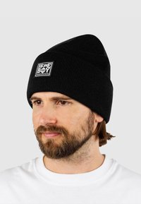 Čierna pletená čiapka s pruhovanou textúrou a prehnutým okrajom. Má štvorcový biely nášivku s logom "HOME BOY loudcouture".