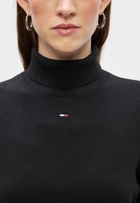 Sweater de gola alta preto feito de tecido tricotado, apresentando um pequeno logotipo tricolor no peito e um decote canelado para adicionar textura.