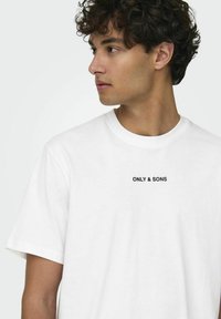 T-shirt bianco in cotone con scollatura a girocollo, con logo stampato nero "ONLY & SONS" al centro. Maniche corte, vestibilità standard.