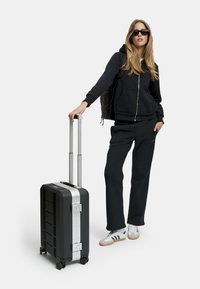 Musta kova kuorimatkalaukku hopeisilla yksityiskohdilla, teleskooppikahva ja pyörät, henkilön vieressä, joka on pukeutunut mustaan huppariin ja verkkareihin.