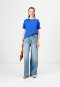 Modellen bär en ljusblå bomullströja och vida, ljusblå jeans, samtidigt som hen håller en brun handväska. Platta, ljusa skor fulländar outfiten.