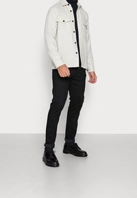 Man i en beige knapp-up jacka över en svart polotröja, svarta uppvikt byxor och svarta läderskor som står på ett ljusgrått golv.