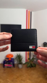 Μαύρο δερμάτινο πορτοφόλι με υφή στο κάτω μέρος, λεία επιφάνεια στο πάνω μέρος και μικρό λογότυπο "TOMMY JEANS" σε κόκκινο, άσπρο και μπλε στο μπροστινό μέρος.