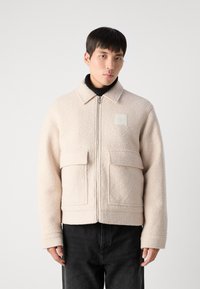 Beige fleece zip-up jacka med krage, två framfickor och en fyrkantig logotypapplikation. Bärs över en svart polotröja och mörka jeans.