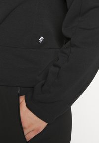 Personne portant un haut noir à manches longues avec un petit logo floral blanc près de la taille, main partiellement dans la poche d'un pantalon noir.