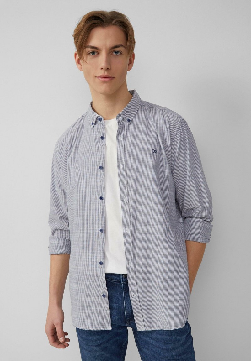 Een getailleerd overhemd in blauw en witte strepen, met lange mouwen, een kraag en een klein logo op de borst; gecombineerd met een wit T-shirt eronder.