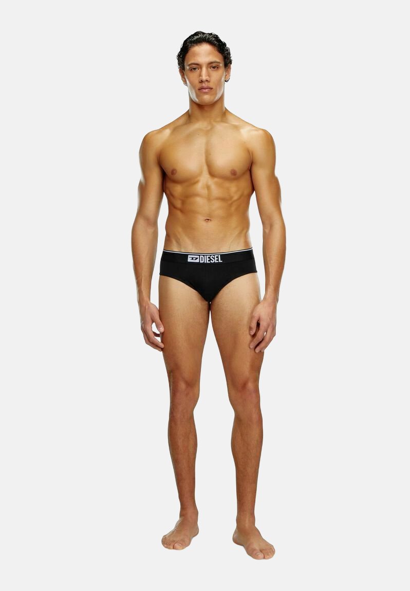 Schwarze Baumwollunterhosen mit einem weißen elastischen Bund, der ein Logo zeigt. Das Model hat einen trainierten Körper und steht barfuß vor einem schlichten Hintergrund.