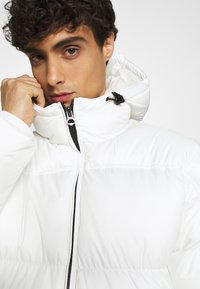 Giacca imbottita bianca con colletto alto, chiusura frontale con zip e dettagli neri. Il materiale sembra liscio, con un design leggero e isolato.