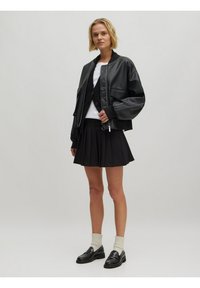 Zwarte leren bomberjack met oversized mouwen, gecombineerd met een witte top en een zwarte geplooide rok. Zwarte loafers en crèmekleurige sokken maken de outfit compleet.