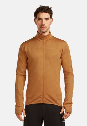 Mężczyzna z krótkimi kręconymi włosami, ubrany w dopasowaną, czerwonobrązową kurtkę na zamek i czarne spodnie, na jednolitym białym tle.