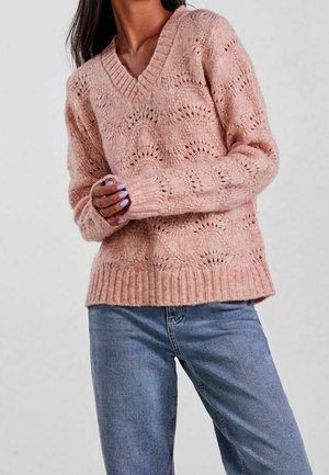 Kobieta w jasno-różowym dzianinowym swetrze z dekoltem w serek i wzorami przypominającymi koronkę oraz niebieskich dżinsach, z rękami złożonymi przed sobą.