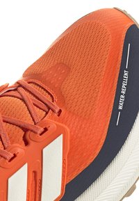 Scarpa sportiva arancione con parte superiore in tessuto testurizzato, punta rinforzata e accenti bianchi. Presenta un'etichetta idrorepellente sul lato.
