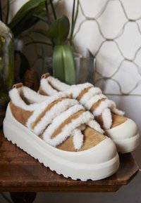 Tan suède sneakers met witte faux-bont banden, dikke witte zool en gestructureerde neuskap, geplaatst op een houten oppervlak met groen in de achtergrond.