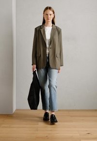 Oliwkowy żakiet na białej fakturowanej bluzce, połączony z jasnoniebieskimi prostymi dżinsami i czarnymi loafersami. Nosi czarną torebkę.