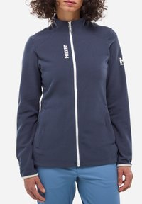 Veste en polaire zippée bleu foncé avec un tissu texturé, fermeture éclair blanche et logo sur la poitrine gauche et la manche. Comprend des poches latérales et un col haut.