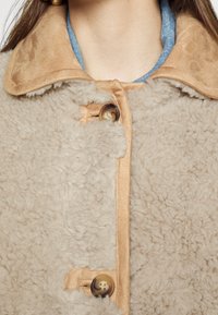 Manteau en shearling beige avec une texture douce, col en satin marron et boutons brun clair. Dispose d'une fermeture à boutons et d'un design décontracté.