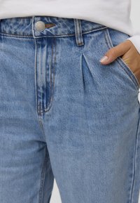 Světle modré džíny z denimu s skládací přední částí, knoflíkovým zapínáním a prošívanými detaily. Tkanina má strukturovaný, vybledlý vzhled.