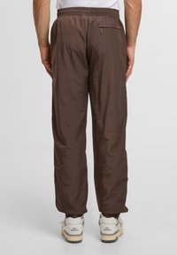 Brązowe, lekkie spodnie z elastycznym pasem, z tylną kieszenią i nieco pomarszczoną teksturą. Ubrane z szarymi i beżowymi trampkami.