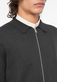 Casaco cinza escuro com fecho zip e tecido texturizado, colarinho pontiagudo e colarinho interior em tom creme. Apresenta um fecho metálico na parte da frente.