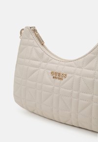 Borsa a tracolla crema trapuntata con hardware dorato e chiusura con zip. Presenta una texture liscia con motivi a rombo. Logo esposto in modo evidente.