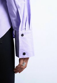 Hånd med sølvring hvilende ved siden, iført lavendelfarvet langærmet skjorte med sorte knapper på manchetten, mod hvid baggrund.