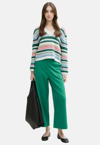 Meerkleurige gebreide trui met horizontale strepen, V-halsontwerp. Groene wijde broek. Zwarte tote bag en platte schoenen.
