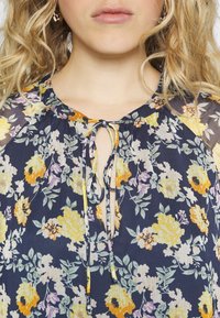 Květinová blůza v námořnické modři se žlutými, fialovými a zelenými vzory. Má kulatý výstřih s detailem na tkaničku v přední části a průhledné rukávy.