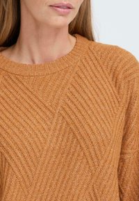 Pull en tricot couleur rouille avec un design texturé en côtes, présentant des motifs diagonaux et un col rond. Matière douce et chaude.