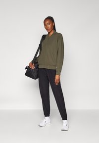 Olivgrüner Kapuzenpullover mit Reißverschluss und gerippten Bündchen, kombiniert mit schwarzen Jogginghosen und weißen Sportschuhen; schwarze Tasche über der Schulter.