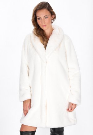 Donna con capelli lunghi che indossa un cappotto in pelliccia sintetica bianca e stivali neri al ginocchio, in piedi contro uno sfondo bianco semplice.