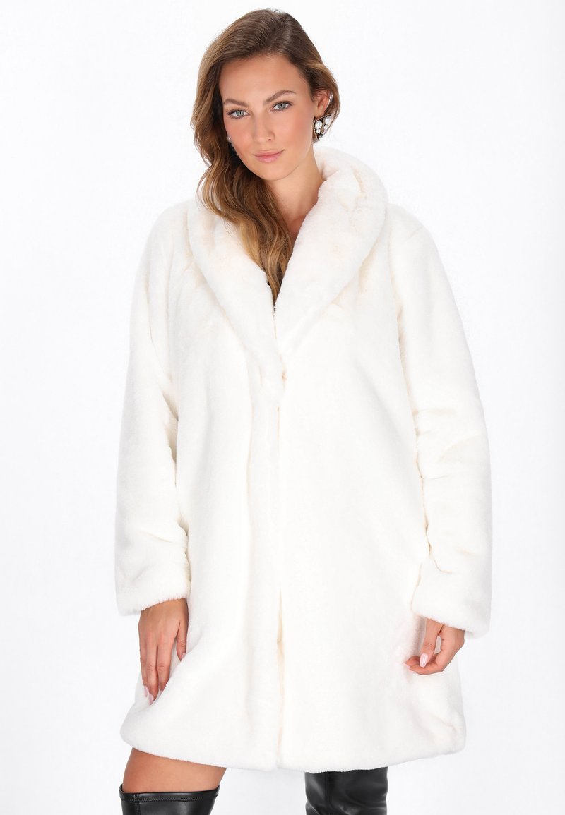 Donna con capelli lunghi che indossa un cappotto in pelliccia sintetica bianca e stivali neri al ginocchio, in piedi contro uno sfondo bianco semplice.