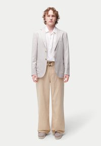 Blazer gris clair avec revers crantés, boutons marron, porté sur une chemise blanche. Pantalon beige à jambes larges et chaussures grises sans lacets. Fond uni.