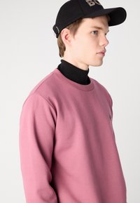 Felpa rosa con scollo rotondo, caratterizzata da un piccolo logo sul petto e indossata sopra un dolcevita nero. Il tessuto appare liscio.