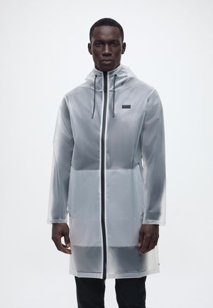 Bărbat purtând o pelerină de ploaie gri translucidă până la genunchi, cu glugă, fermoar frontal și șnururi negre, stând pe un fundal simplu.