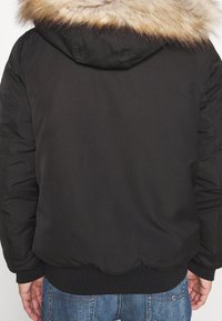 Bakifrån av en person som bär en svart hujacka med beige pälskanter och blå denimjeans. Händer syns vid sidorna.