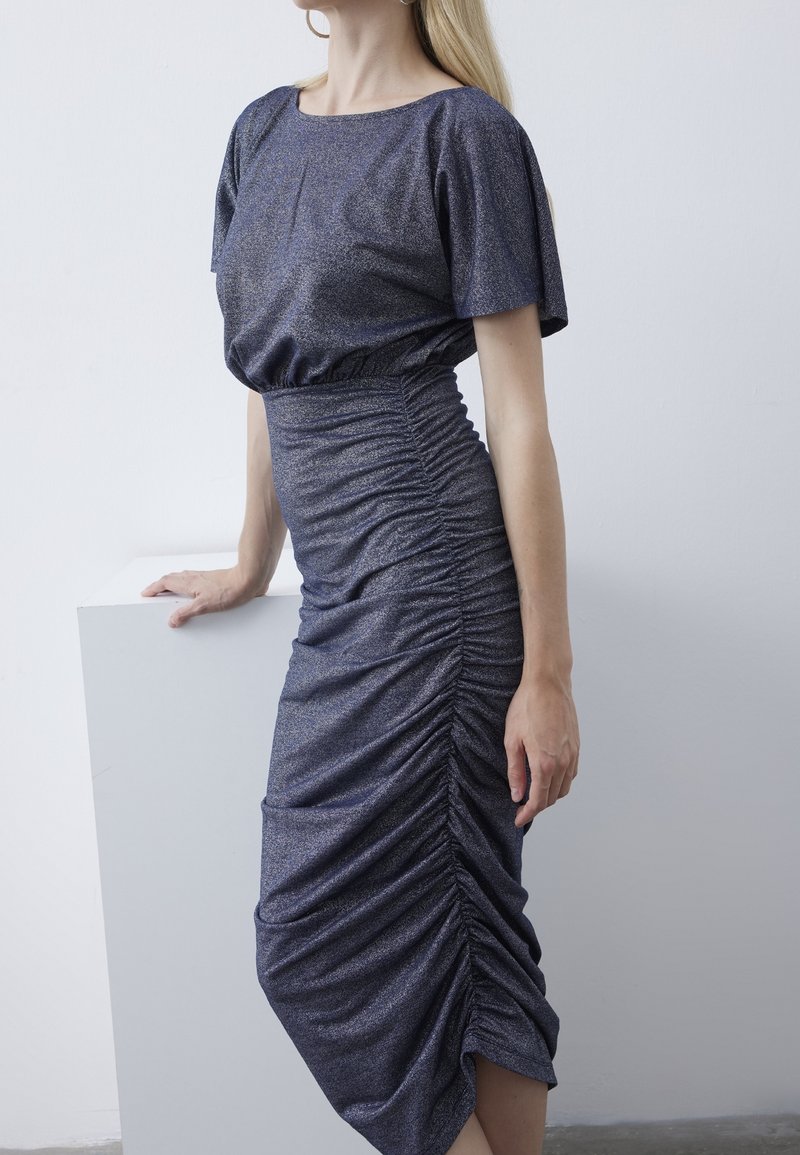 Robe bleu marine froncée en tissu brillant et texturé. Elle présente des manches courtes et une silhouette ajustée, créant un effet effilé à l'ourlet.