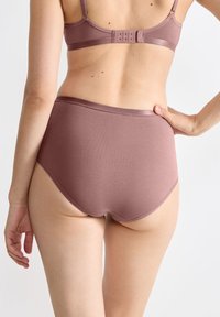 Vrouw gezien van achteren, gekleed in mauve katoenen hoge taille ondergoed en bijpassende beha met haak-en-oogsluiting, hand op de heup.
