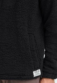 Sudadera de forro polar sherpa negra con una superficie texturizada, que cuenta con un bolsillo lateral y una etiqueta de tela blanca con el texto de la marca.