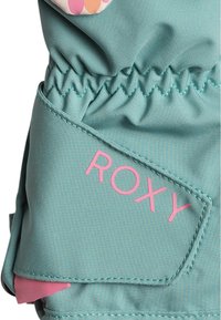 Tissu vert avec une texture lisse, mettant en avant un logo "ROXY" rose sur une pièce triangulaire ; l'élastique froncé en haut assure un ajustement confortable.