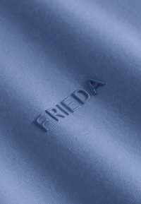 Blauwe stof met een gladde textuur, voorzien van verhoogde letters die "FRIEDA" in een modern lettertype lezen. Minimalistisch ontwerp, geen extra patronen.