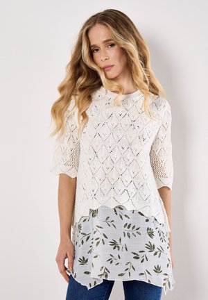Lace-knit white top with half sleeves, layered over a light, patterned blouse featuring green leaf accents, worn with blue jeans.