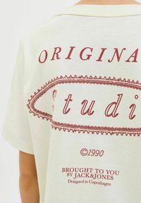 Kreminių spalvos marškinėliai su trumpomis rankovėmis, featuring bordo spalvos grafine tekstine dizainu, kuris skaito "ORIGINAL Studio" ir "Atneša jums JACK&JONES."