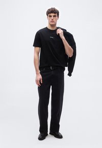 Νέος άνδρας που φοράει μαύρο μπλουζάκι, μαύρα τζιν και μαύρα παπούτσια, κρατώντας ένα μαύρο σακάκι στον ώμο του μπροστά σε απλό φόντο.