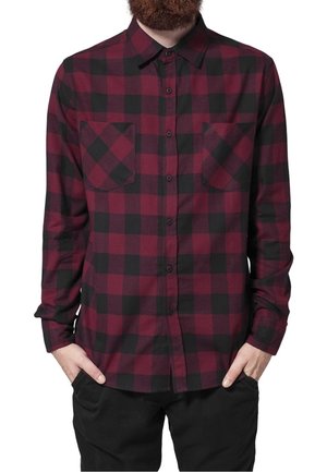 Homme portant une chemise à manches longues rouge et noire à carreaux avec deux poches poitrine et un pantalon noir, les mains dans les poches.