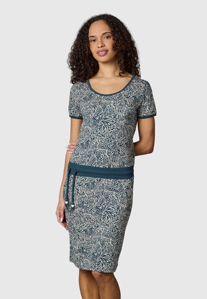 Kvinde med krøllet hår iført en blå og hvid blomsterprintet kortærmet kjole med snøre i taljen, stående foran en ensfarvet baggrund.